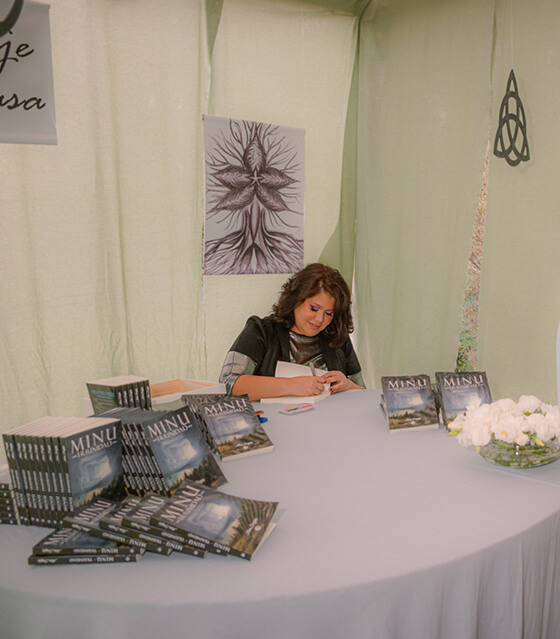 Autora Monidragon firmando un libro en stand lleno de libros